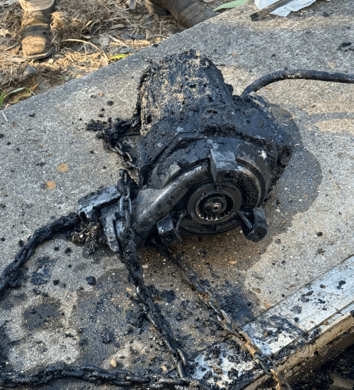 Burnt submersible pump covered in sludge and debris after removal from a wastewater pit, resting on concrete with a damaged power cable and chain.