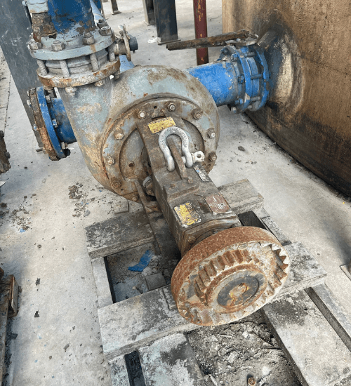 Old industrial pump installed on a concrete pad, showing rusted gears and flanges connected to blue process piping in a plant environment.