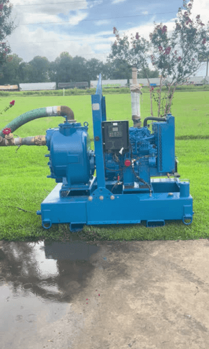 Rebuilt Tier 3 trash pump skid painted blue on a steel frame with forklift pockets. Features discharge piping with butterfly and ball valves, green suction hose, and a diesel engine with control panel. Photographed on concrete in Lafayette, LA.