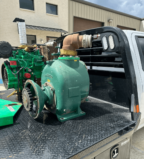 Green industrial diesel pump assembly sitting on a flatbed truck outside a metal building, showing the discharge flange and gear drive for field service or rebuild.