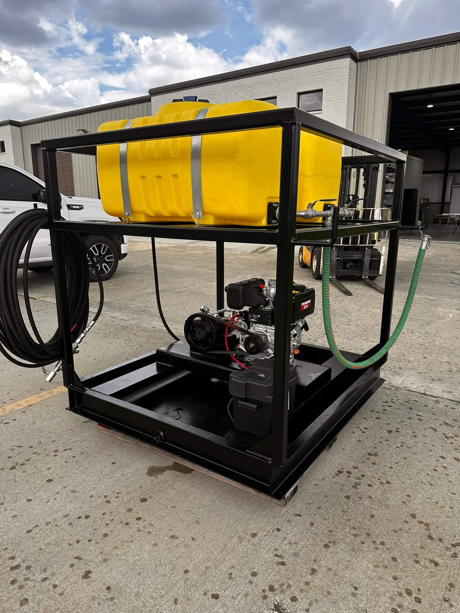 Angled front-right view of the chemical injection skid showing the twin yellow chemical tanks, stainless piping, and large black hose coil mounted on the side, set outdoors on concrete with green grass behind.