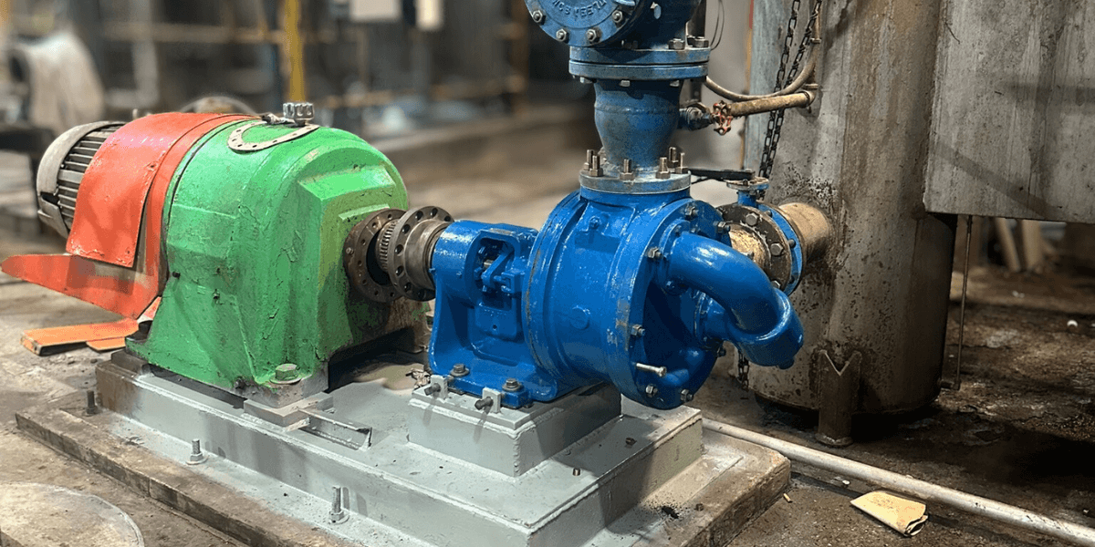 Rebuilt industrial pump system mounted on a newly refurbished skid inside a facility. The blue pump and green motor are bolted to a clean, gray skid, surrounded by piping and equipment in a heavy-duty plant environment.